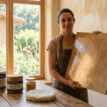 Profesional de la bioconstrucción mostrando panel de estuco de cal pulido con Com Cal Cera Natural junto a envases del producto y trapo de lana.